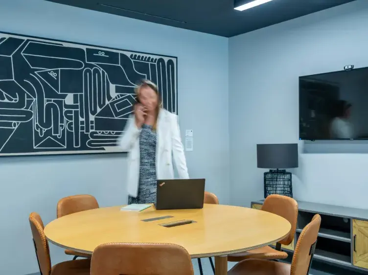 Una mujer de pie hace una llamada telefónica en una sala de reuniones con mesa redonda, sillas marrones, laptop, lámpara, televisión en la pared y una pintura abstracta.