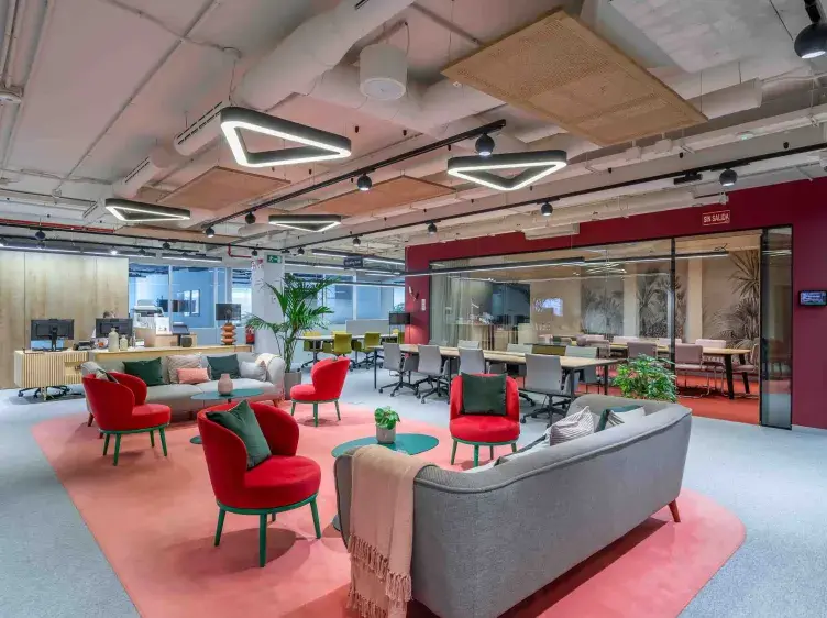 Sofás grises rodeados de sillas rojas en una oficina moderna, con mesas de trabajo y plantas. Letrero: "EN SALA".