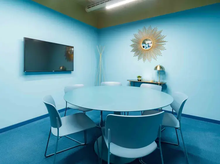Una mesa redonda azul rodeada de sillas está en una sala con paredes y suelo azules. Un televisor cuelga en la pared; un espejo decorativo y una lámpara están al fondo.