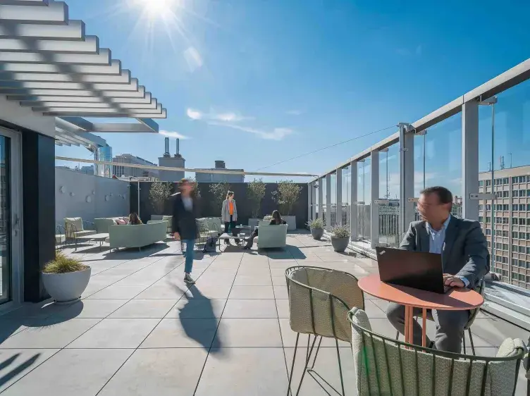 Hombre trabajando en una laptop en una mesa al aire libre. Personas conversan en sillones en una terraza urbana bajo el sol brillante.