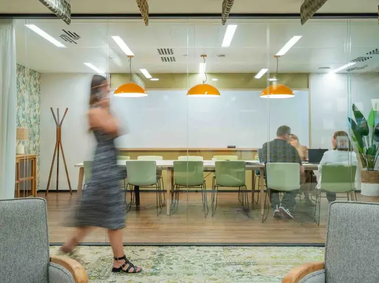 Mujer caminando en oficina mientras tres personas conversan dentro de una sala de reuniones con pared de vidrio.