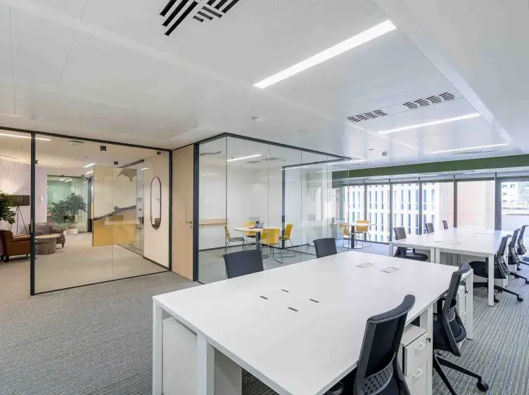 Espacio de oficina moderno; mesas blancas y sillas negras en línea; paredes de vidrio separan salas, con luz natural entrando por ventanas amplias.