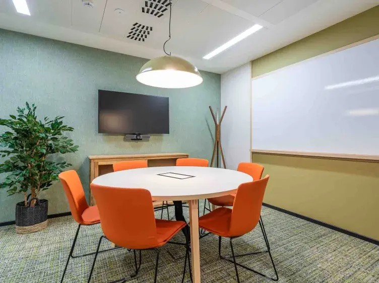 Mesa redonda blanca con cinco sillas naranjas alrededor. Está en una sala de reuniones con televisión, pizarra blanca, planta decorativa, perchero de pie y lámpara colgante.