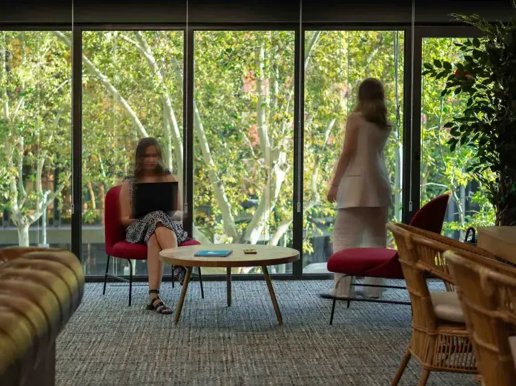 Una persona sentada usa una laptop sobre una mesa en un salón con grandes ventanas mostrando árboles. Otra persona está de pie, de espaldas, mirando hacia afuera.