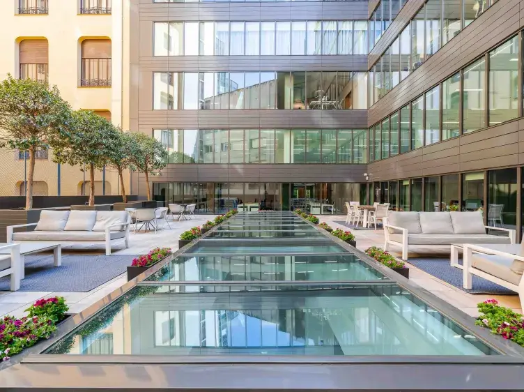 Una terraza moderna con sofás blancos y mesas rodea una piscina estrecha de vidrio. Edificios de oficinas la rodean, con ventanas reflejando el cielo. Plantas decoran los bordes.