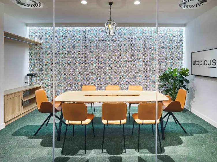 Una mesa rectangular rodeada de sillas naranjas está en una sala de reuniones con pared de mosaico.