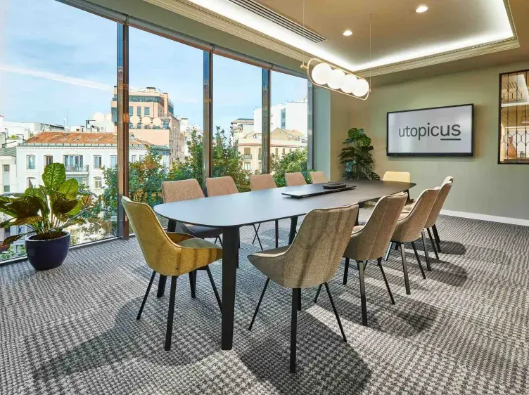 Mesa de conferencia con sillas modernas rodeada de ventanas grandes; en la pared, pantalla muestra "utopicus". Oficina con piso alfombrado y planta decorativa en el rincón.