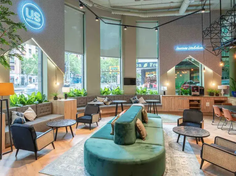 Espacio de descanso con sofás verdes y mesas en entorno moderno. Ventanales grandes, plantas decorativas y luces colgantes.