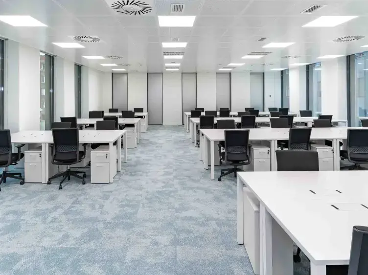 Oficina con escritorios vacíos y sillas negras distribuidos simétricamente; ambiente moderno y bien iluminado con ventanas grandes.