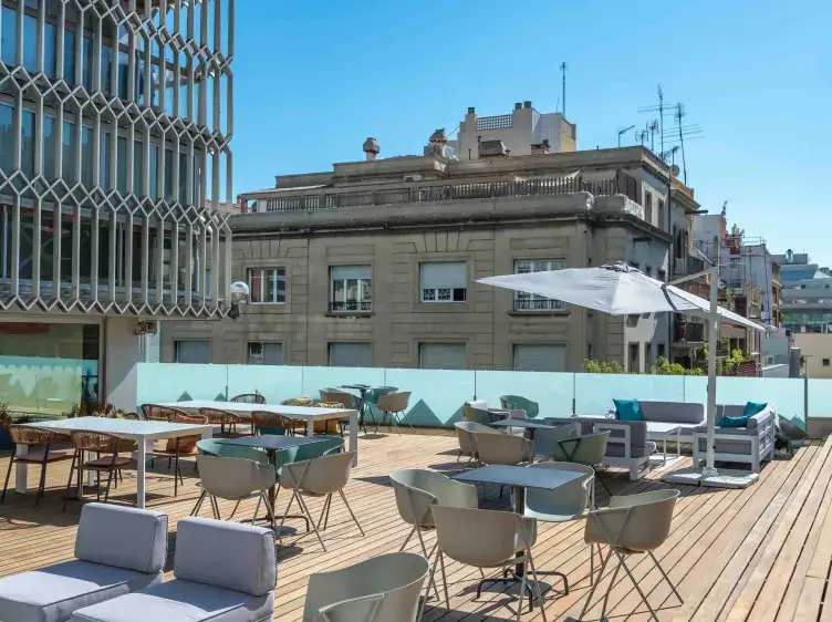 Terraza con mesas y sillas dispersas, junto a sombrillas. Edificios circundantes tienen fachadas grises y ventanas cerradas bajo un cielo despejado.