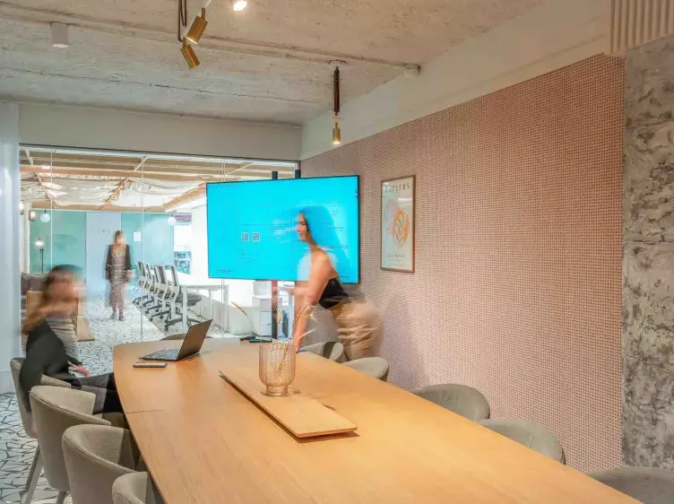 Pantalla mostrando contenido digital en una sala de reuniones moderna con personas interactuando y mesas de madera. Una persona está de pie mientras otra está sentada frente a un portátil.