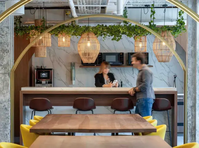 Una mesa de madera rodeada de sillas amarillas se sitúa en un espacio moderno de cocina con lámparas colgantes y vegetación decorando. Al fondo, dos personas interactúan junto a la barra.