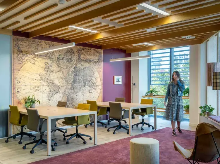 Una sala de reuniones con mesas y sillas, adornada con un mapa antiguo. Una mujer camina hablando por teléfono, junto a una ventana soleada con plantas.
