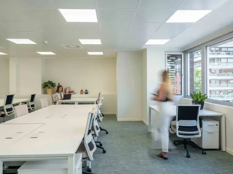 Una oficina bien iluminada muestra escritorios blancos en filas. Una mujer camina mientras otra trabaja en su computadora. Ventanas grandes permiten ver edificios exteriores.