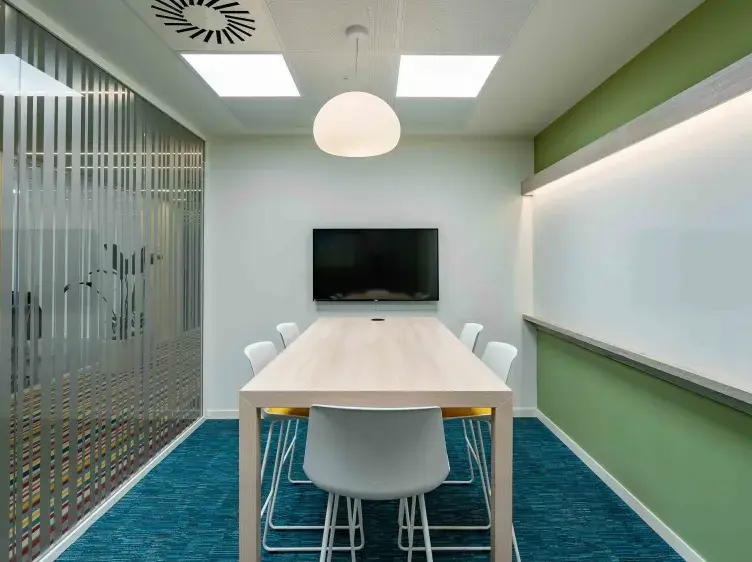 Mesa rectangular con seis sillas blancas, ubicada en una sala de reuniones; pantalla montada en la pared, y pared acristalada a la izquierda. Decoración moderna y minimalista.
