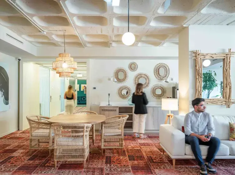 Una sala luminosa con decoración bohemia; un hombre está sentado en un sofá blanco, mientras dos mujeres caminan y observan objetos decorativos en la pared.