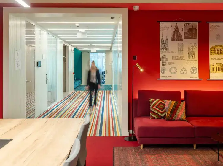 Sofá rojo y mesa de madera en una oficina con paredes rojas. Persona caminando por un pasillo de rayas coloridas. Decoración con lámpara y cuadros informativos.
