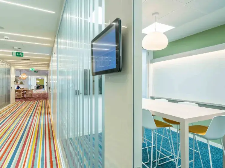 Una pantalla está montada en la pared de un pasillo con alfombra de rayas. A la derecha, una sala de reuniones moderna con una mesa blanca y sillas.