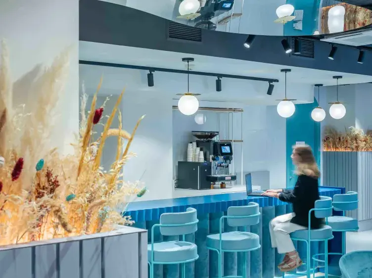Una mujer trabaja en una computadora portátil en un bar moderno de tonos azules, rodeada de decoración floral y lámparas colgantes.