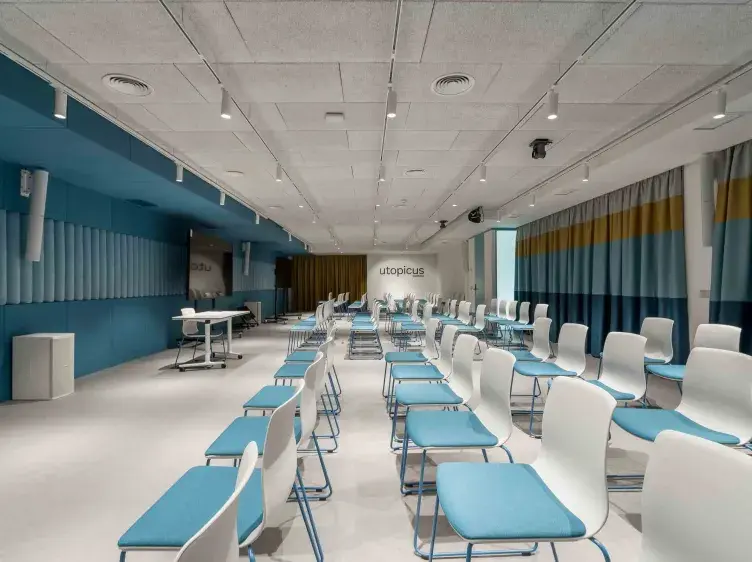 Filas de sillas blancas con asientos azules, dispuestas en una sala amplia y moderna; al fondo, el texto "utopicus" en la pared.
