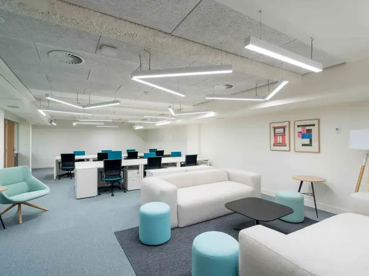Sala de oficina moderna; sofás blancos y mesas bajas azules en primer plano; escritorios con computadoras y sillas en el fondo. Decorado con arte en las paredes.