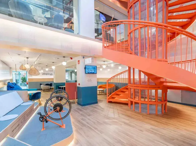 Escalera de caracol naranja sube en oficina moderna; a la izquierda, exhibición de bicicleta, sofás azules. En la pared se lee "utopicus" junto a pantallas y lámparas colgantes.