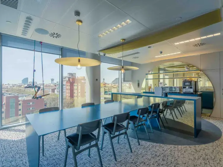 Mesa alargada con sillas alrededor, bajo luces colgantes. Ambiente moderno de oficina con ventanales y área de descanso. Al fondo, una vista de edificios urbanos.