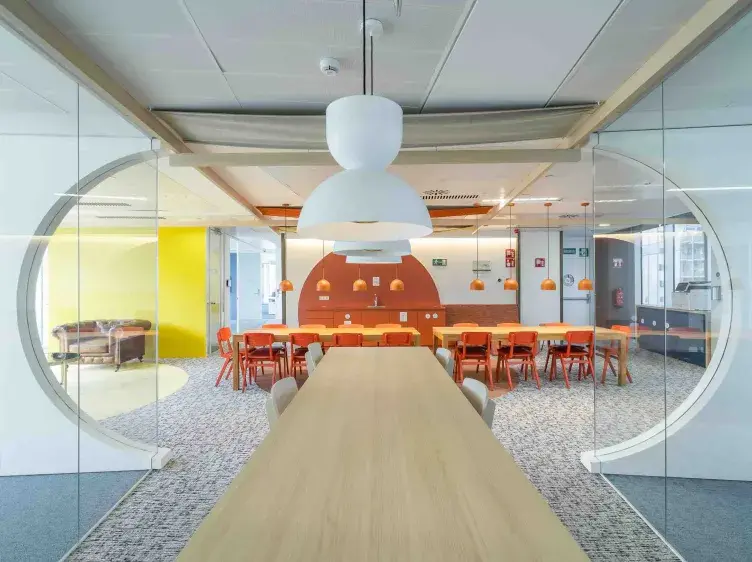 Mesa larga de reuniones, situada bajo lámparas blancas colgantes, en una sala con paredes de vidrio, sillas naranjas y fondo con pared amarillo claro.