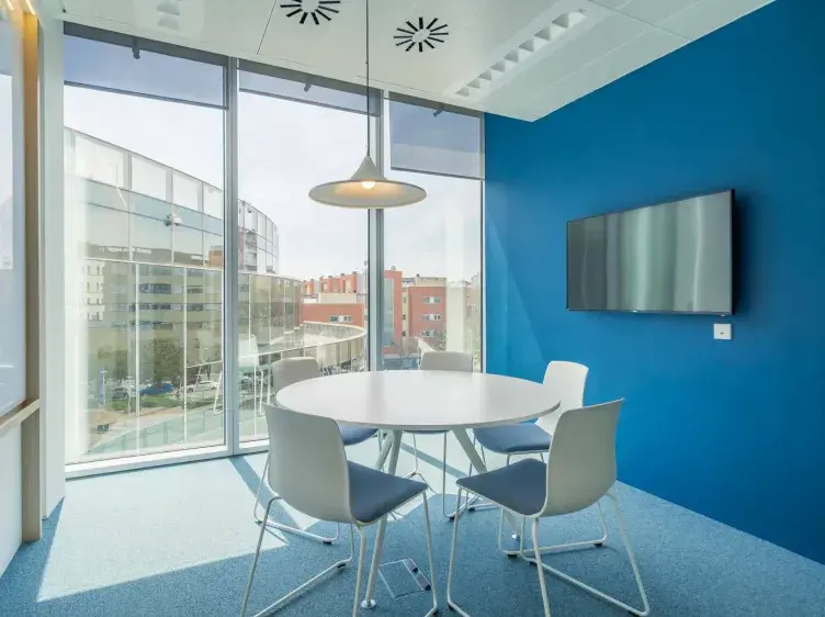 Una mesa redonda con cuatro sillas, ubicada en una sala de reuniones pequeña con paredes azules. Una pantalla plana está montada en la pared, junto a grandes ventanas con vista a edificios.