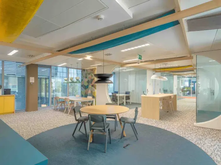 Mesa redonda con sillas alrededor, en un espacio de oficina moderno con paredes de vidrio y decoración colorida. Ventanas amplias permiten la entrada de luz natural al ambiente.