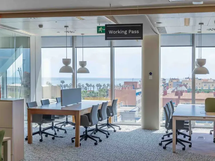 Mesas y sillas de oficina, dispuestas para trabajo, en una sala con ventanas grandes. Letrero: "Working Pass". Al fondo, vista de edificios y el mar.