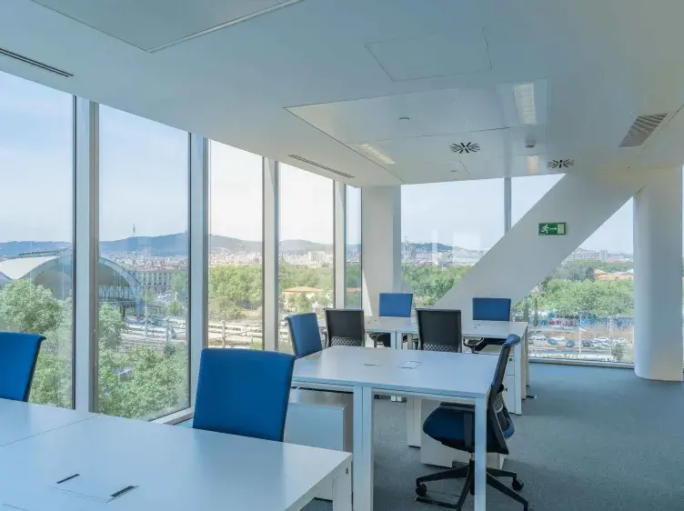 Oficina moderna con escritorios blancos y sillas azules, vista a través de grandes ventanas hacia una ciudad y paisaje montañoso. Señal verde de salida de emergencia en la pared.