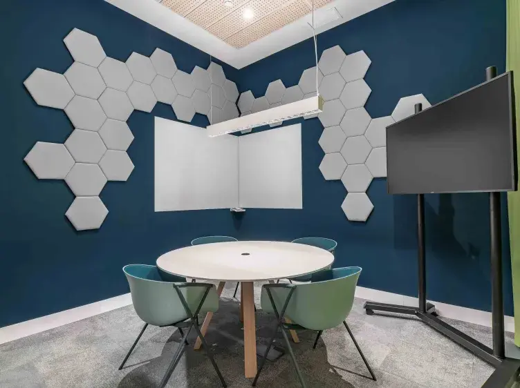 Mesa redonda rodeada de cuatro sillas en sala de reuniones. Paredes con paneles hexagonales, pizarra blanca y pantalla sobre soporte.