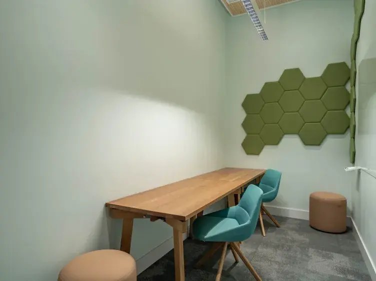 Mesa de madera alargada con dos sillas azules, ubicada en una sala de paredes verdes con paneles hexagonales decorativos. Un puf marrón complementa el mobiliario en un espacio moderno.