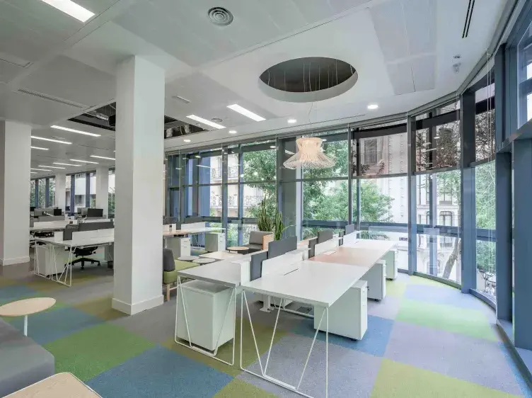 Espacio de oficina moderno con escritorios blancos, sillas ergonómicas y mamparas divisorias. Ventanas grandes permiten la entrada de luz natural; plantas decoran el ambiente.