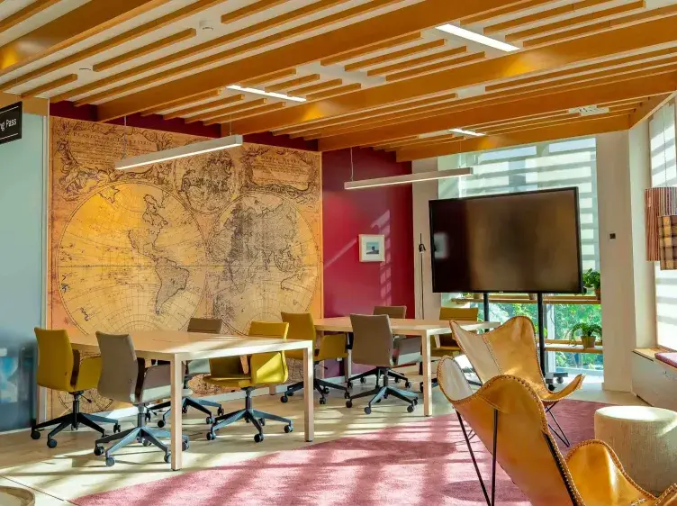Espacio de trabajo con mesas y sillas sobre ruedas, mapa antiguo en la pared, pantalla grande, iluminación moderna y ventanales que permiten la entrada de luz natural.