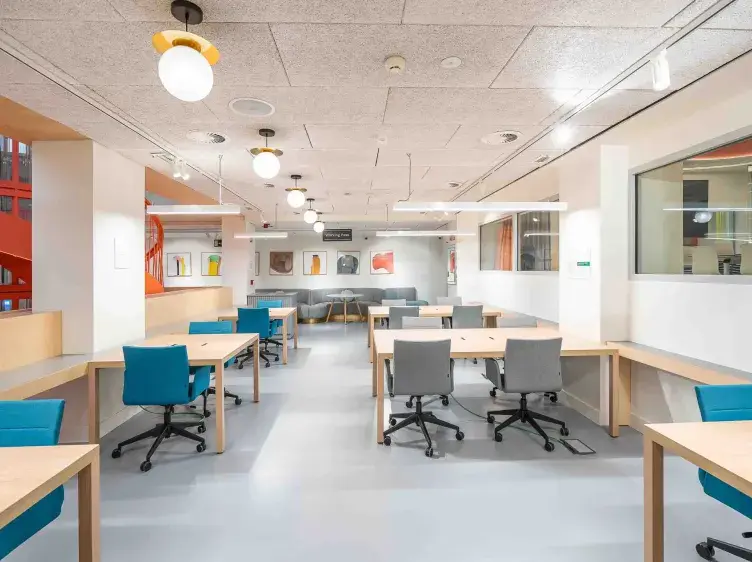 Espacio de oficina con escritorios de madera y sillas giratorias de colores, iluminado por lámparas modernas, rodeado de paredes blancas decoradas con cuadros abstractos.