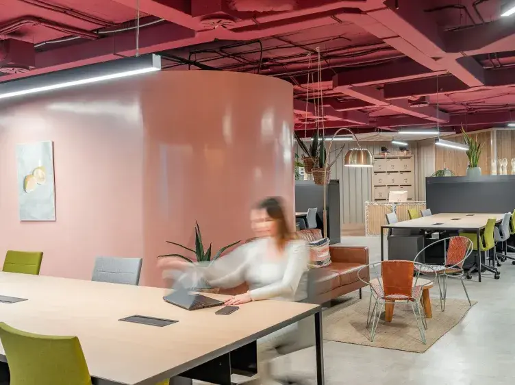 Persona sentada moviéndose frente a una mesa con laptop, rodeada de sillas y decoración moderna en una oficina de techo rosa.
