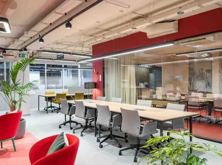 Una mesa larga con sillas giratorias en una oficina moderna; plantas y mobiliario colorido rodean el espacio.