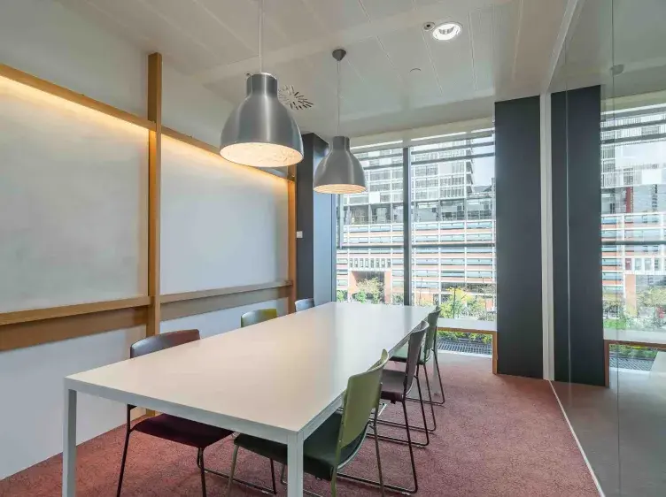 Una mesa blanca rectangular con sillas multicolores está desocupada en una sala de reuniones con grandes ventanales y lámparas colgantes modernas, iluminando el espacio.