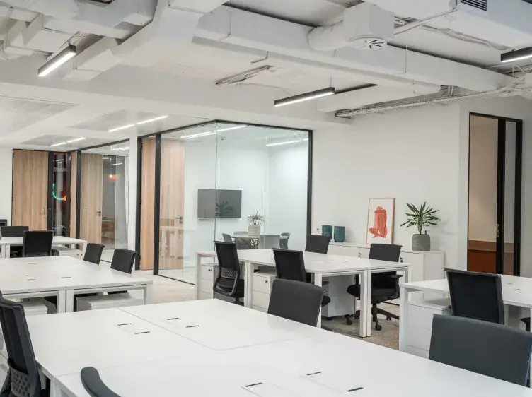 Mesas blancas ordenadas con sillas negras en una oficina moderna, con paredes de cristal y decoración minimalista.