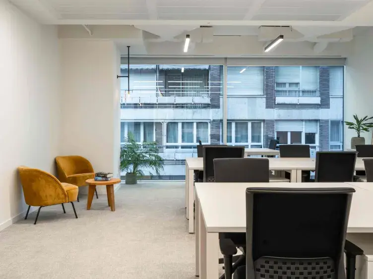 Muebles de oficina están dispuestos en una sala luminosa con ventanas grandes; sillones amarillos y plantas añaden calidez al ambiente moderno.