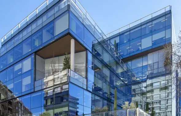 Un edificio moderno con fachada de vidrio refleja el cielo y sus alrededores; plantas decoran varias terrazas.