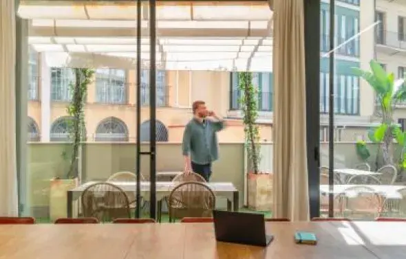 Una sala de reuniones con mesa de madera y sillas. Un hombre de pie habla por teléfono en un balcón, rodeado de edificios y plantas.