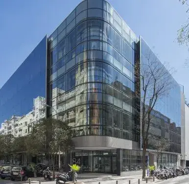 Un edificio moderno de cristal refleja la luz del sol. Coches estacionados en la calle a su alrededor, en un entorno urbano con árboles y construcciones vecinas.