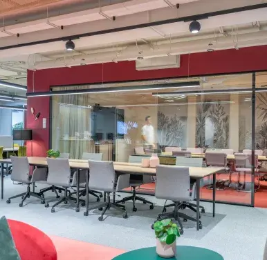 Un espacio moderno con sillones rojos y grises, personas conversan y trabajan en un entorno luminoso. Decoración contemporánea y plantas. Letreros visibles: "SALIDA" y un panel con pantalla iluminada.