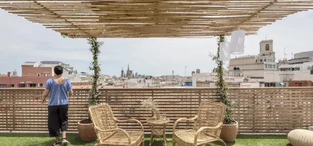 Una persona observa desde una terraza con pérgola de bambú y muebles de mimbre. Se ve una ciudad al fondo bajo un cielo despejado.