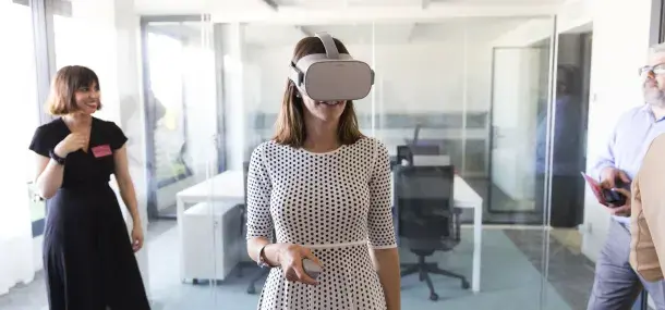 Una mujer usa gafas de realidad virtual en una oficina moderna con paredes de vidrio, mientras otras personas observan.