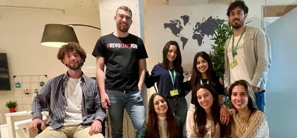 Un grupo de ocho personas sonríe a la cámara; algunos están sentados y otros de pie. Detrás hay un mapa del mundo en una pared y una planta. Una persona viste una camiseta con el texto "#REVOLUTION".