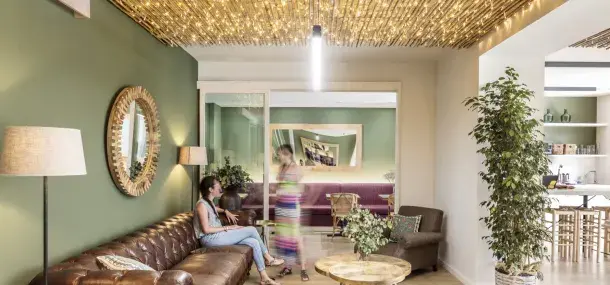 Una persona sentada en un sofá marrón interactúa con otra de pie frente a un espejo decorativo. La habitación tiene paredes verdes, iluminación cálida y plantas decorativas.